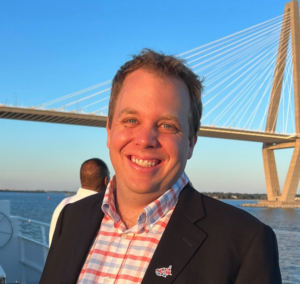 A headshot of a blonde-haired man wearing a blue and red checkered shirt, he is smiling in front of river with a bridge.