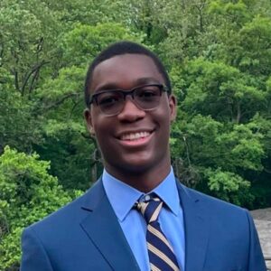 A headshot of a smiling, college-aged Black man in glasses wearing a blue suit and a blue and gold striped tie.