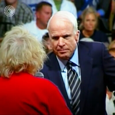 An older man in a suit talks to a woman in red holding a microphone in a crowded indoor setting.