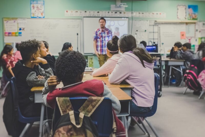 A classroom with students sitting at tables facing a teacher who is standing and talking at the front near a whiteboard.