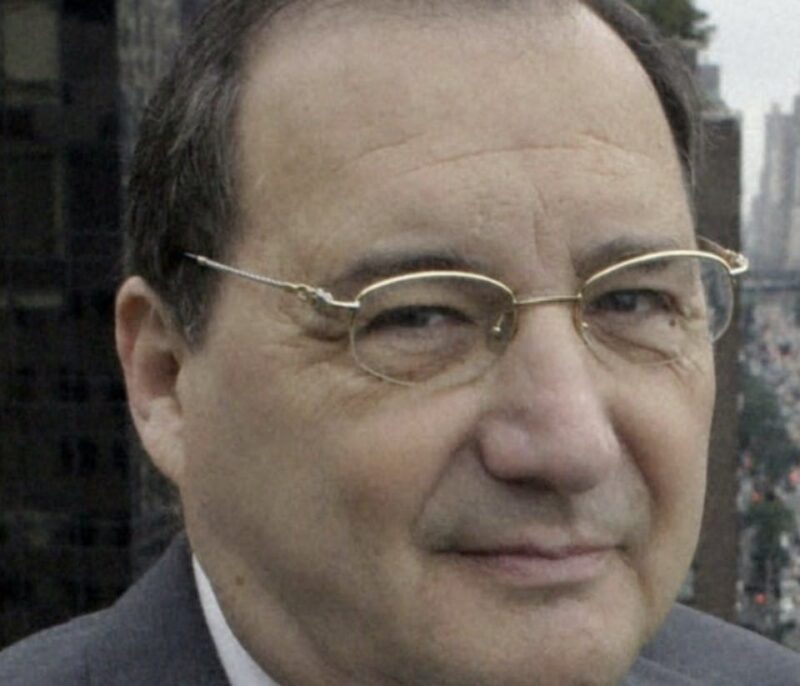 Man with short hair, glasses, and a suit, standing outdoors with a cityscape in the background.