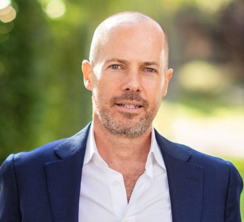 A man with a shaved head and beard wearing a white shirt and blue blazer, standing outdoors with a blurred green background.