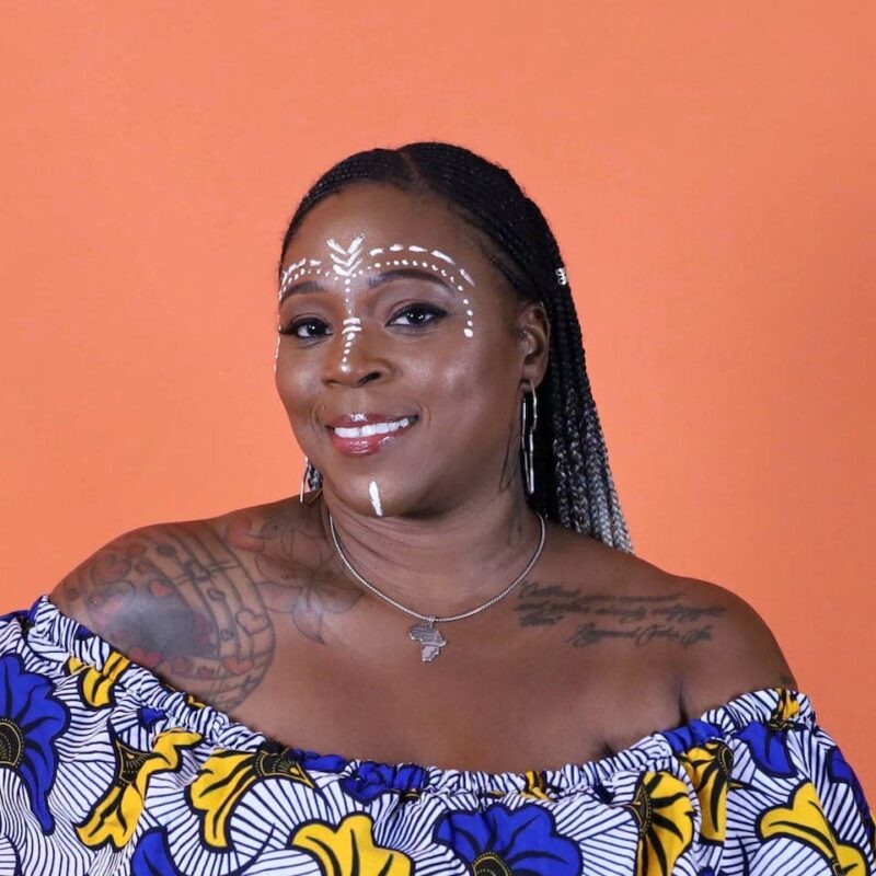 Smiling person with face paint, braided hair, floral top, and visible tattoos, stands against an orange background.