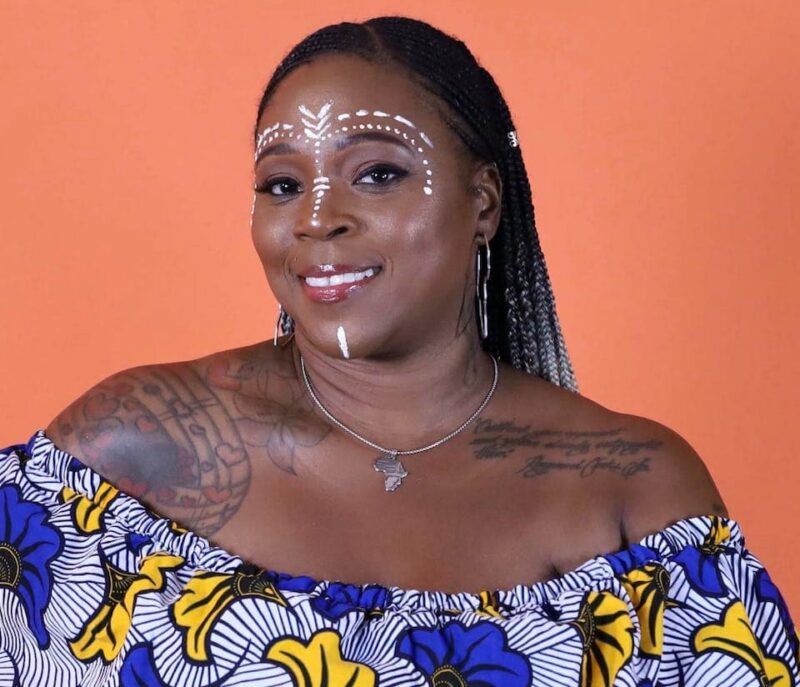 Smiling person with face paint, braided hair, floral top, and visible tattoos, stands against an orange background.