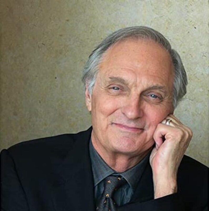 An older person with gray hair smiles, resting their chin on their hand. They wear a black suit and tie, against a beige background.