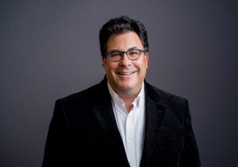 Smiling man in a dark blazer and white shirt against a gray background.