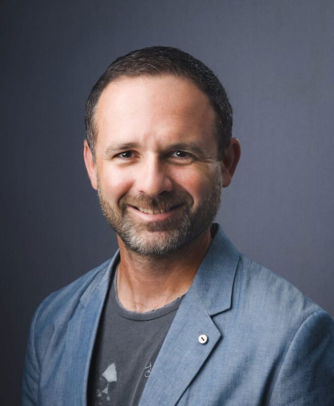 Smiling man with short dark hair and beard, wearing a gray blazer and t-shirt, against a plain dark background.