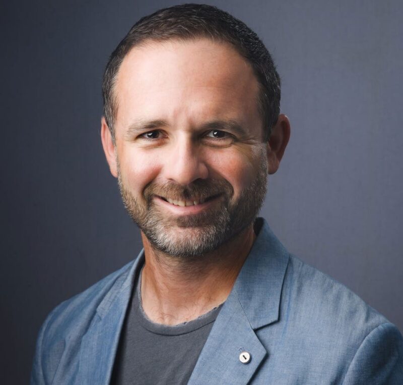 Smiling man with short dark hair and beard, wearing a gray blazer and t-shirt, against a plain dark background.