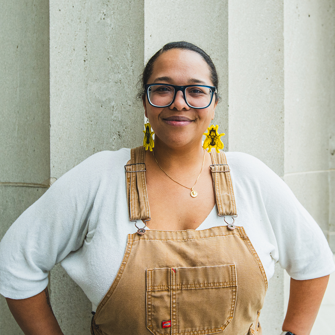 Person smiling, wearing glasses, sunflower earrings, white shirt, and brown overalls, standing against a concrete pillar.