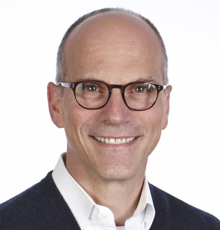 A smiling man wearing glasses and a dark sweater over a white shirt against a plain background.