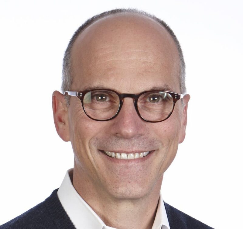 A smiling man wearing glasses and a dark sweater over a white shirt against a plain background.