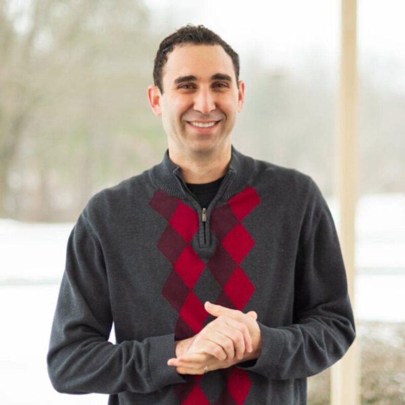 Smiling person wearing a dark sweater with red diamond pattern, standing outdoors on a snowy day.