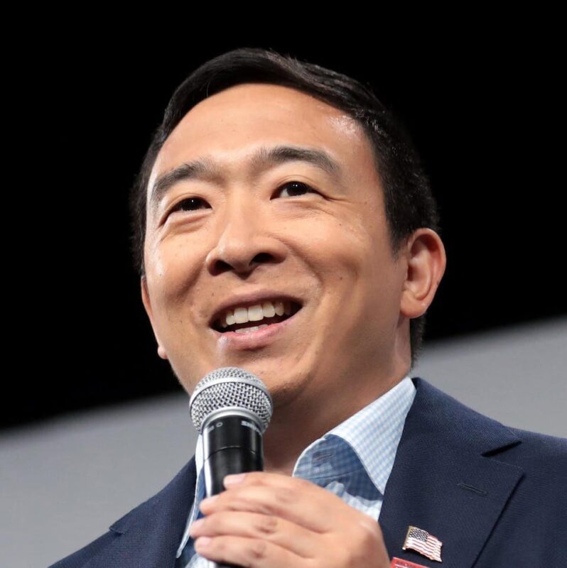 A person smiling while holding a microphone, wearing a suit with an American flag pin.