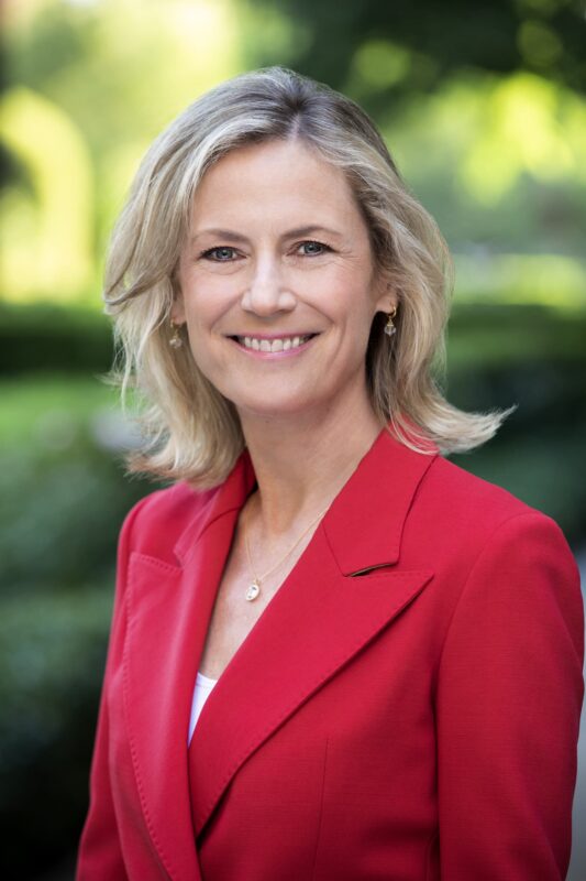 Smiling woman with shoulder-length blonde hair in a red blazer stands outdoors with leafy background.