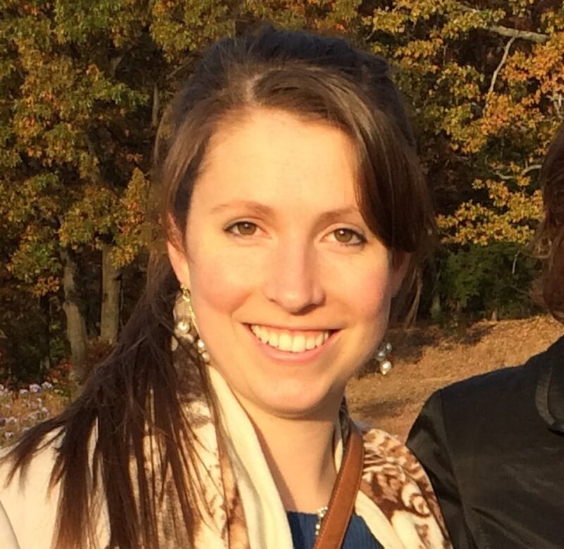 Woman smiling outdoors with autumn trees in the background.