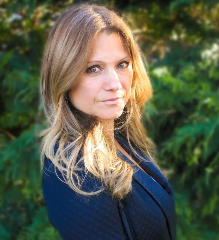 Woman with long, light brown hair wearing a dark top, standing outdoors with greenery in the background, looking at the camera.