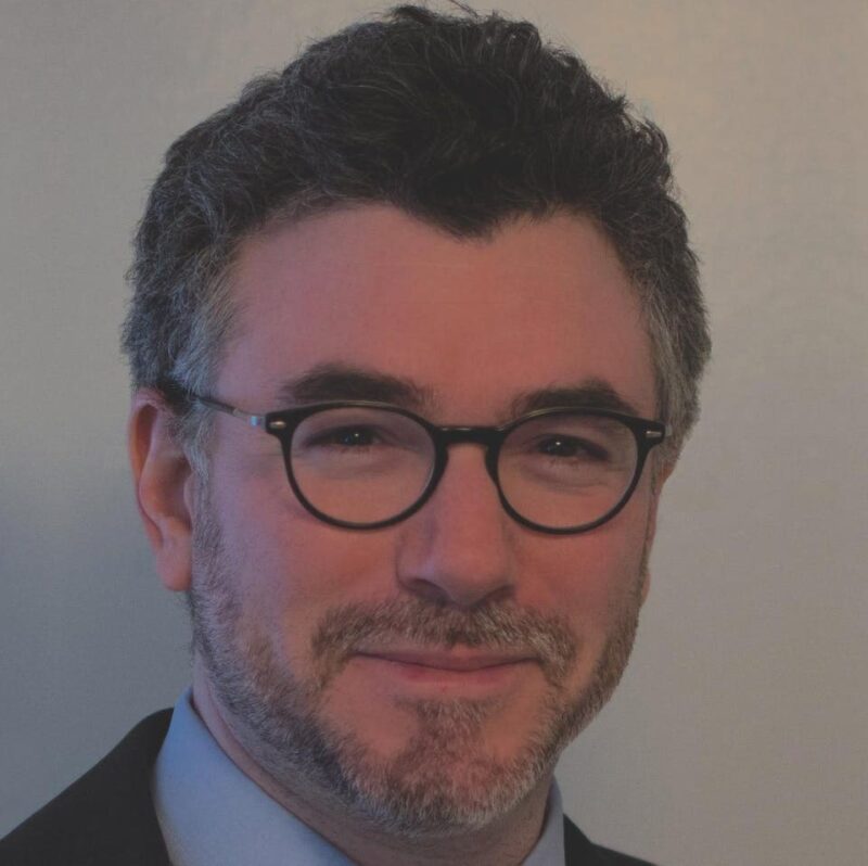 A person with short, curly hair, wearing glasses and a suit, smiles at the camera.