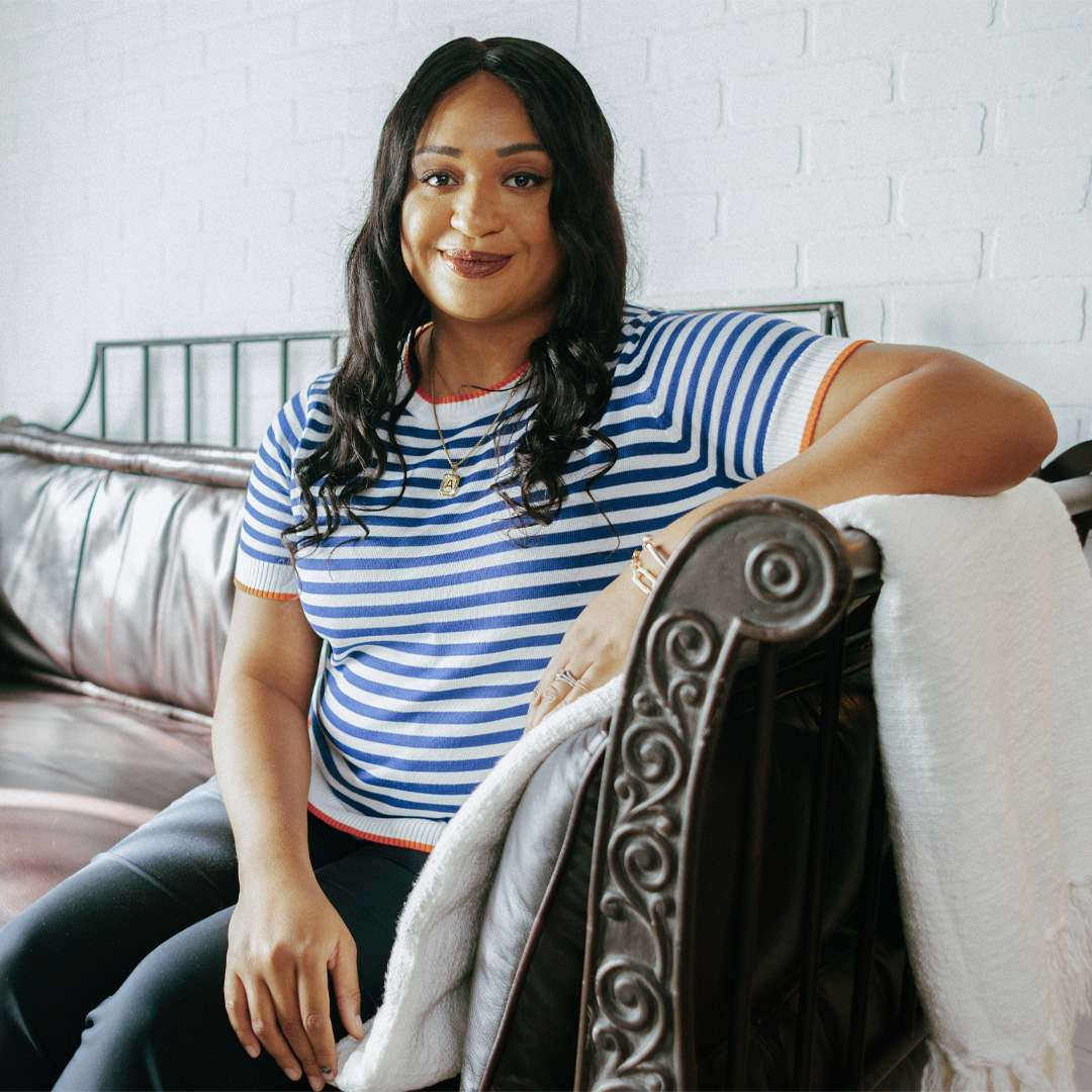 Person in a striped shirt smiles, sitting on a brown leather sofa with a white blanket, against a brick wall backdrop.