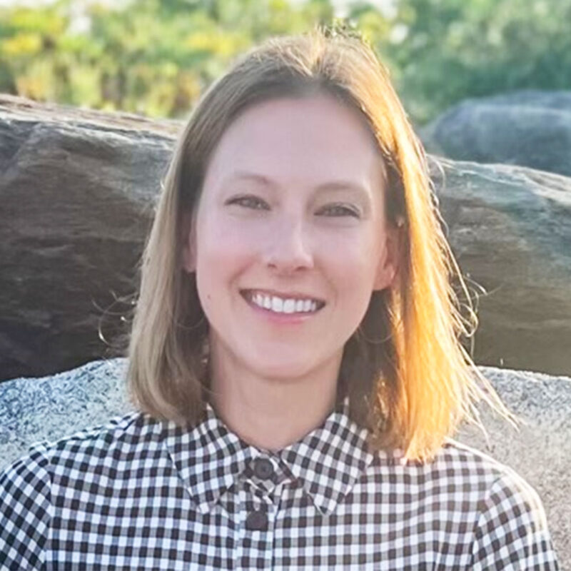 A person with short hair smiles, wearing a checkered shirt, posing outdoors with rocks and greenery in the background.