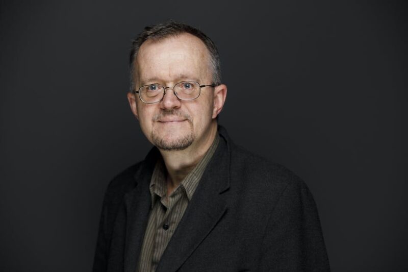 Man with glasses and beard wearing a dark blazer and striped shirt, standing against a dark gray background.