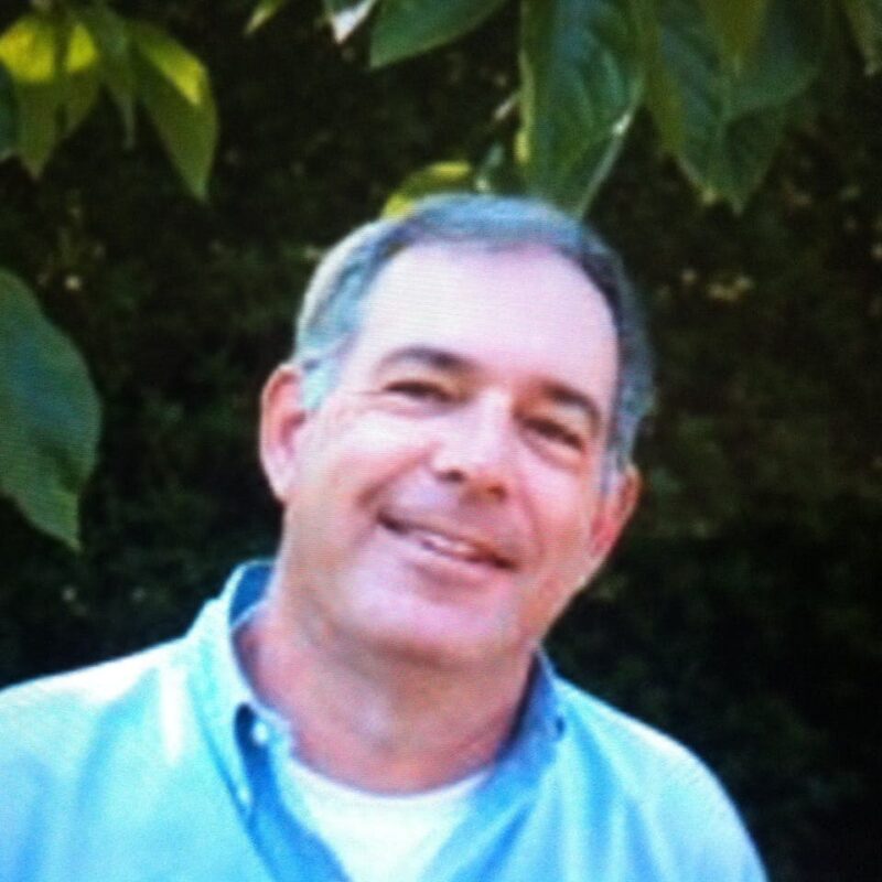 A man in a light blue shirt smiles outdoors, standing in front of greenery and leaves.