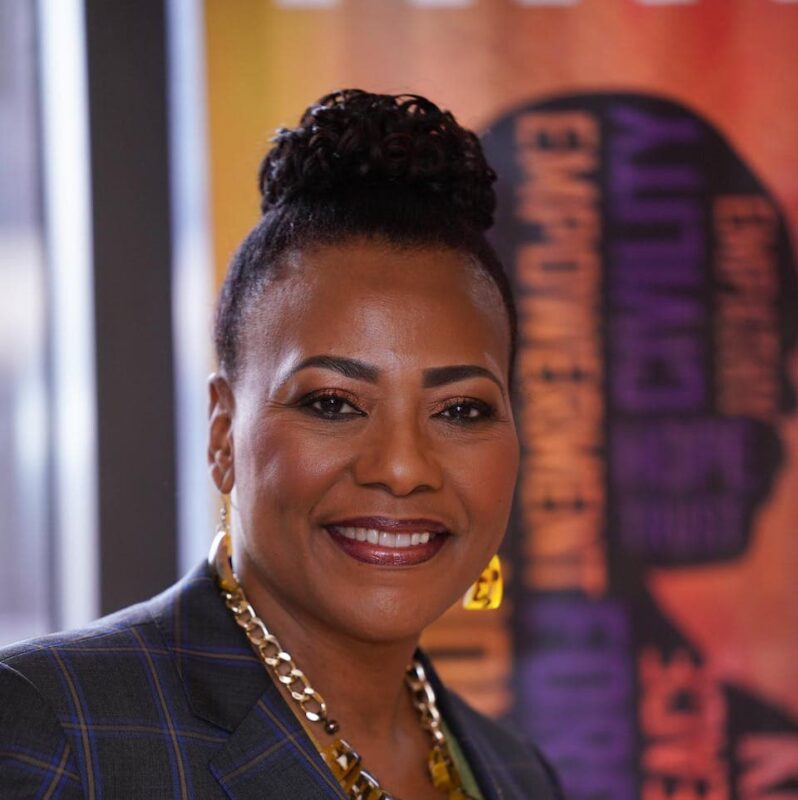 Person with braided bun, earrings, and necklace smiling in front of a colorful, blurred background with text.