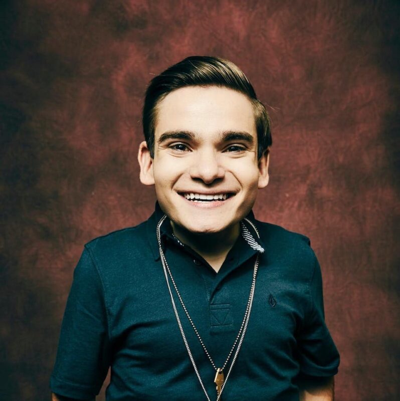 Person with a big smile wearing a dark polo shirt and necklace against a maroon textured background.