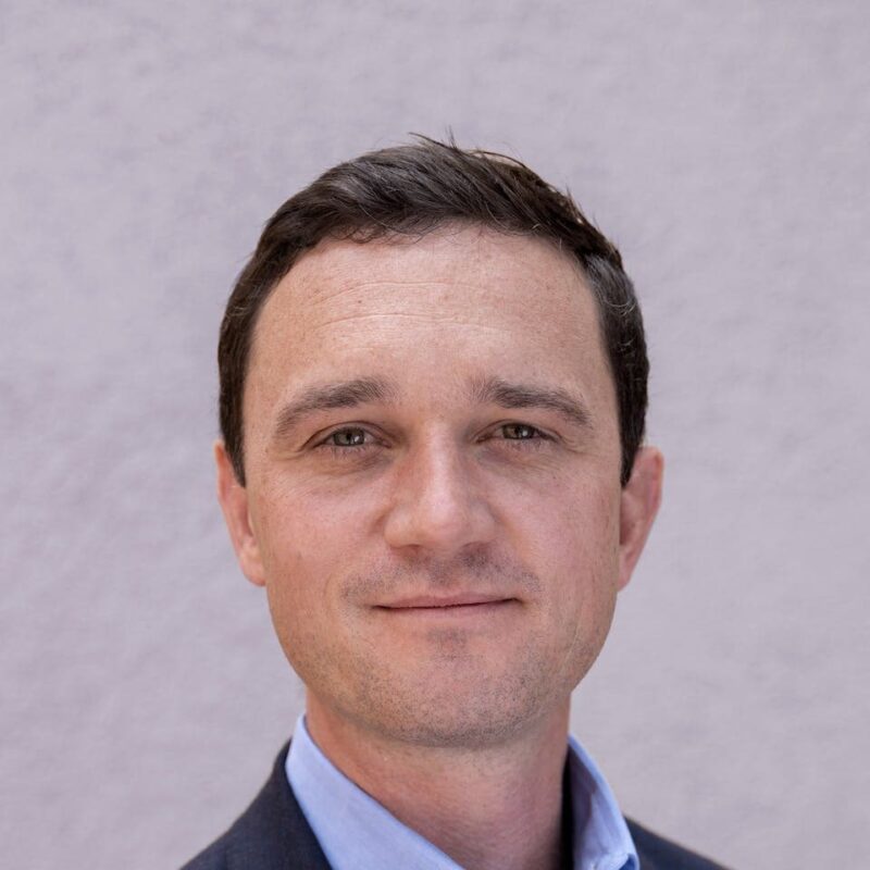 A man in a suit, with short dark hair, looking at the camera, against a plain light gray background.