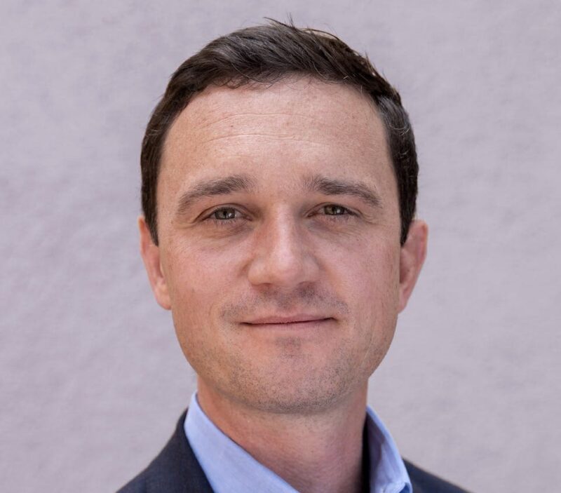 A man in a suit, with short dark hair, looking at the camera, against a plain light gray background.
