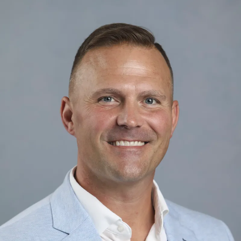 Smiling man in a light gray suit jacket and white shirt against a plain gray background.