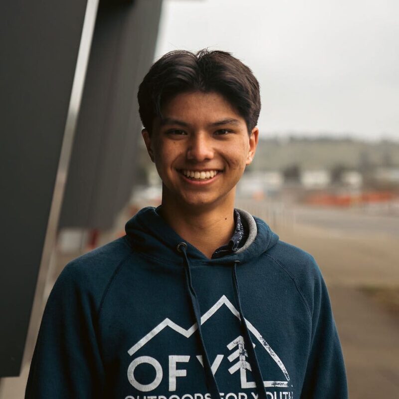 A person smiling, wearing a dark hoodie with a mountain design, stands outdoors with a blurred background of buildings and hills.