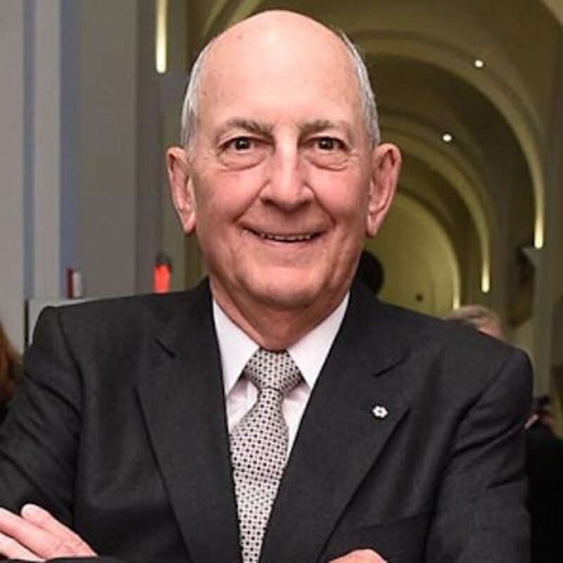 Bald man in a suit smiling with arms crossed, standing indoors with a softly lit archway in the background.