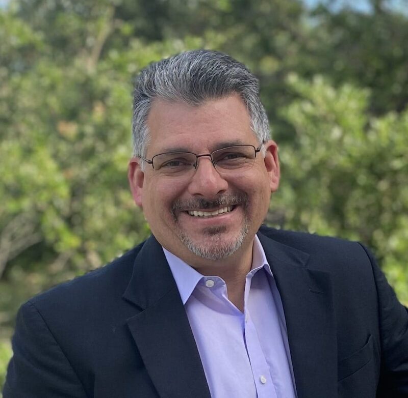 Smiling person wearing a dark suit and glasses, standing outdoors with greenery in the background.