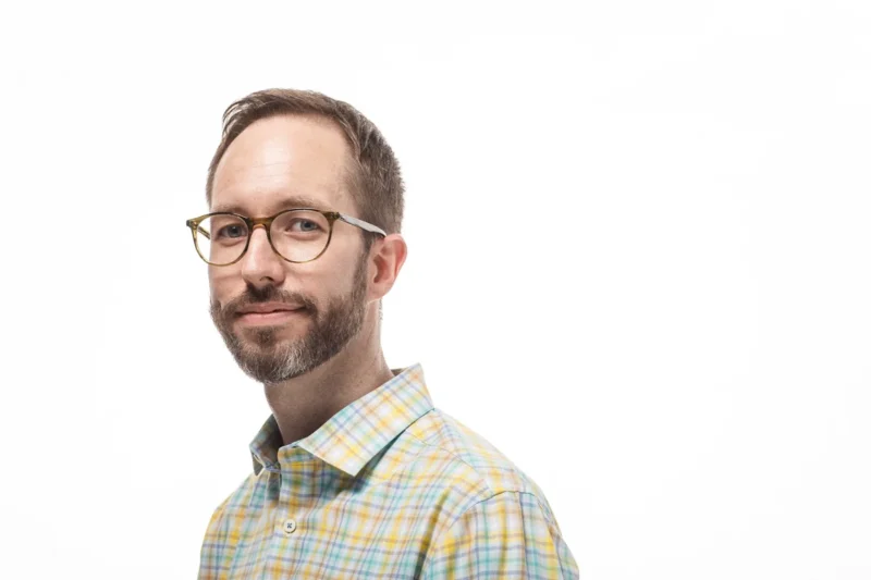 Man with glasses, beard, wearing a plaid shirt, looking at the camera against a white background.