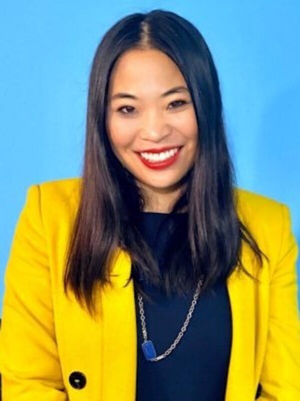 Smiling woman with long black hair wears a yellow blazer over a black top, against a blue background.