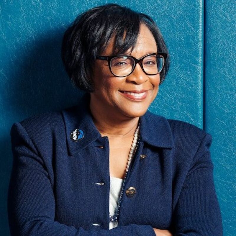 Smiling woman with short dark hair and glasses, wearing a navy blazer, stands against a blue textured wall.