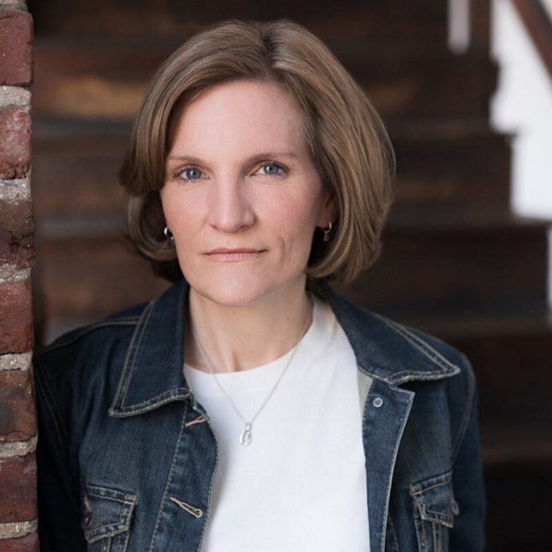 Person with short hair wearing a denim jacket and white shirt, standing in front of a brick wall and staircase.