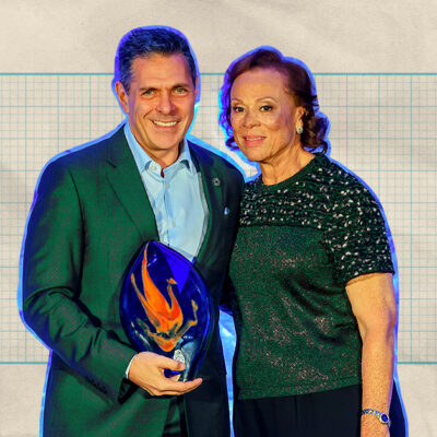 A man in a suit holding a glass award stands beside a woman in a sequined top against a graph paper background.