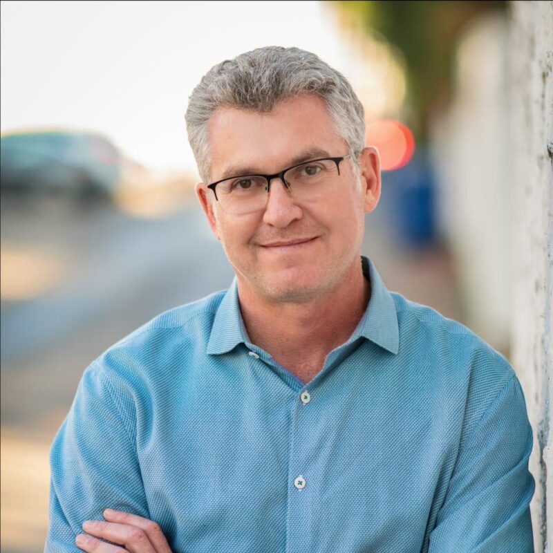 Man with short gray hair and glasses, wearing a blue shirt, standing outdoors with blurred background.