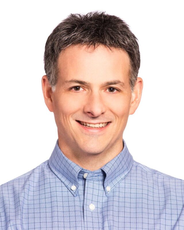 Smiling person with short hair wearing a light blue checkered shirt against a plain white background.