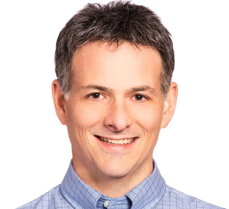 Smiling person with short hair wearing a light blue checkered shirt against a plain white background.