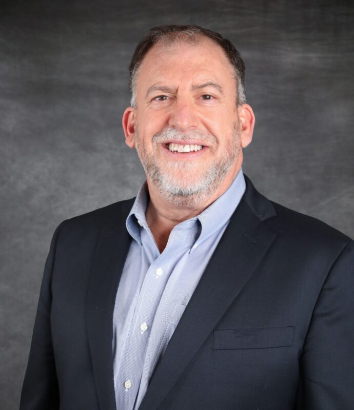 Smiling man in a dark suit and light shirt poses against a gray textured backdrop.