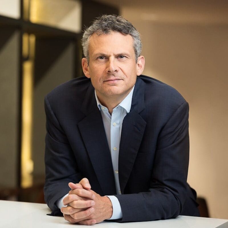 A man in a dark suit and light shirt leans forward on a white table, hands clasped, with a calm expression. Neutral background.