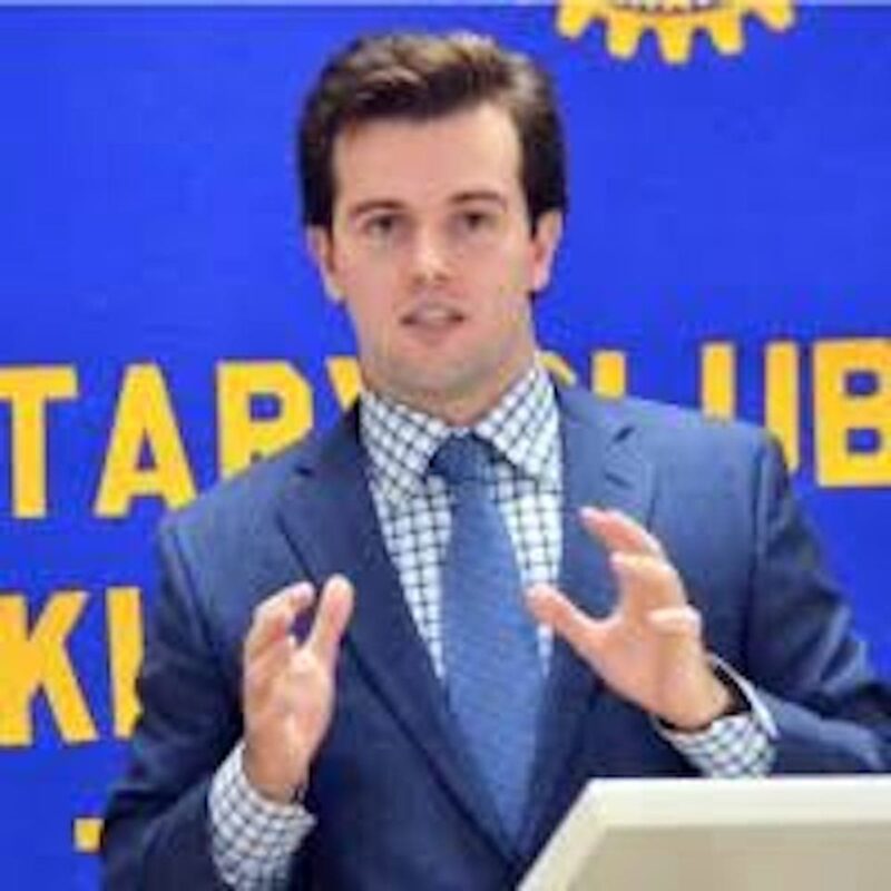 A man in a suit and tie speaks at a podium, gesturing with his hands. A blue and yellow Rotary Club banner hangs in the background.