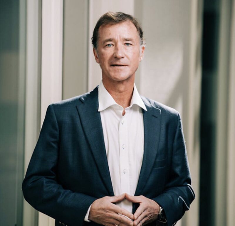 Man in a navy blazer and white shirt stands indoors with hands clasped, softly smiling.