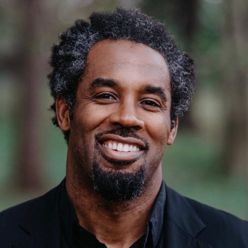 Smiling man with curly hair and a beard, wearing a dark jacket, standing outdoors with blurred greenery in the background.