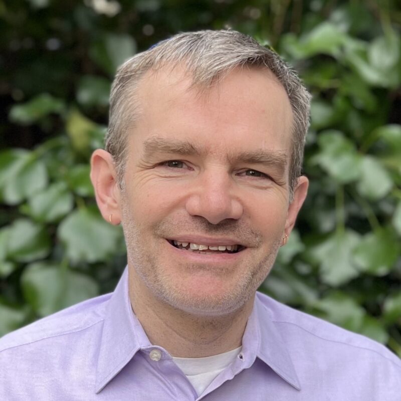 A person with short gray hair smiling, wearing a light purple shirt, standing in front of a background with green leaves.