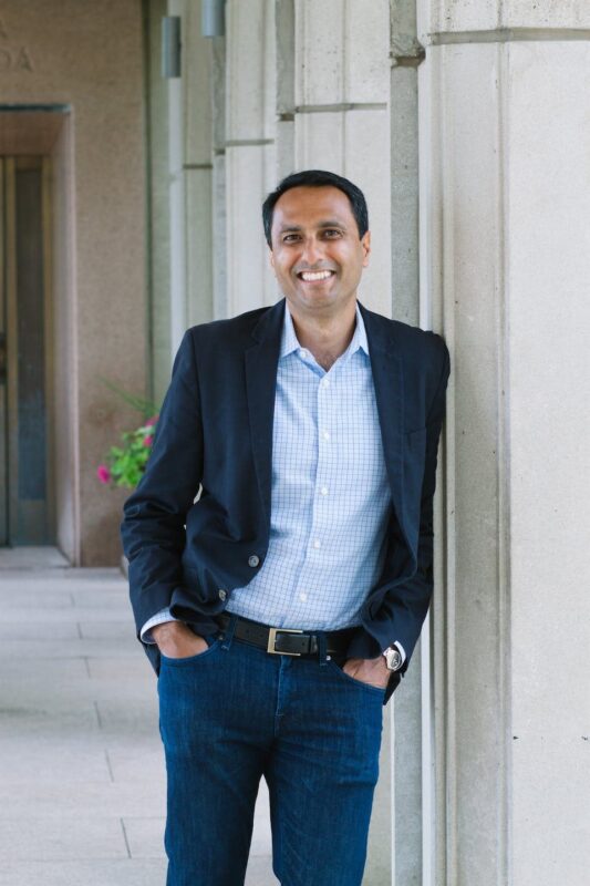 A man in a blazer and jeans leans against a column in a corridor, smiling with hands in pockets.