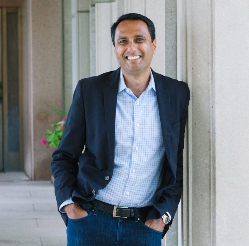 A man in a blazer and jeans leans against a column in a corridor, smiling with hands in pockets.