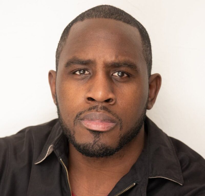 Man with short hair, beard, and serious expression wearing a black jacket over a red shirt, against a plain white background.